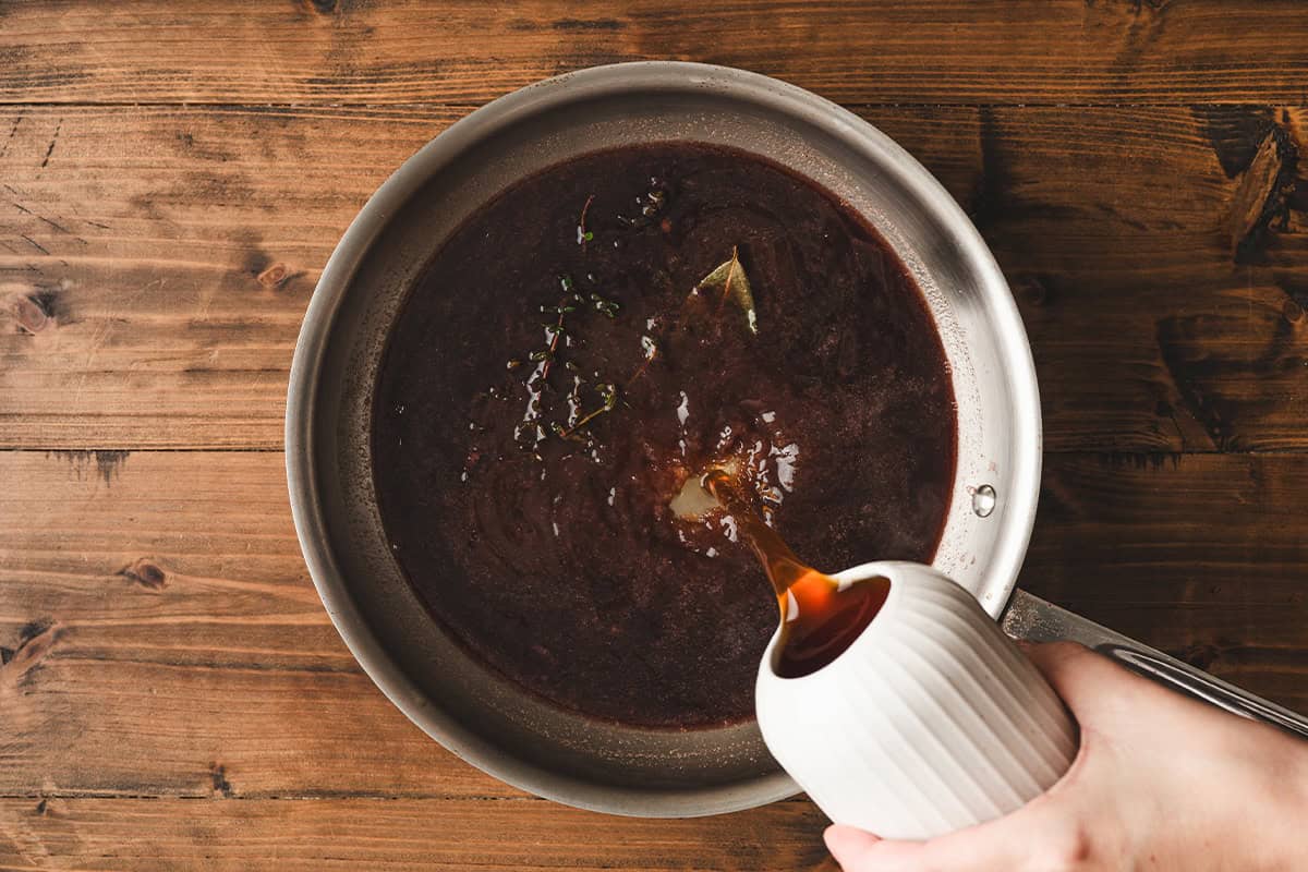 Beef stock being poured into the simmering red wine and herb mixture in a stainless steel skillet.