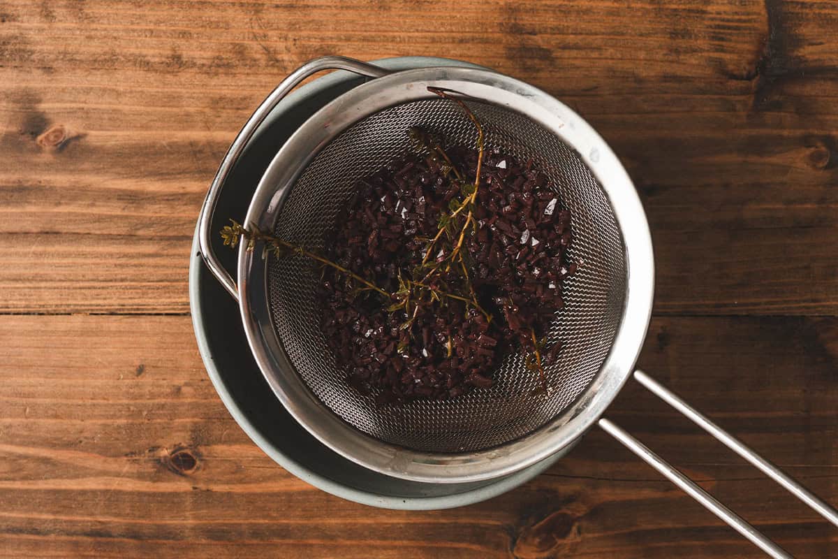 Straining the reduced red wine sauce through a fine mesh sieve to remove the cooked shallots, garlic, and herbs.