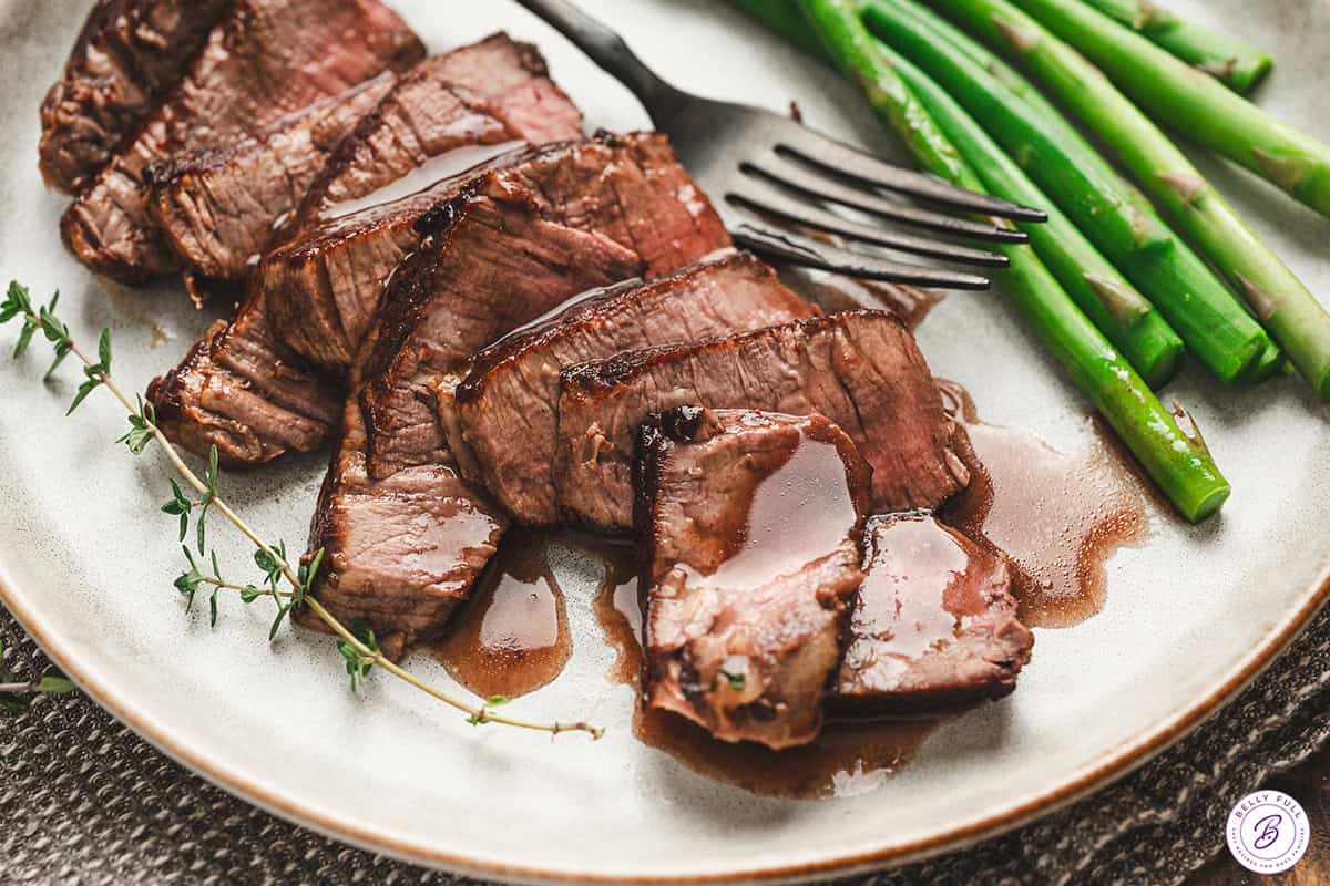 Close-up of sliced steak drizzled with homemade red wine sauce, served with fresh thyme and bright green asparagus.