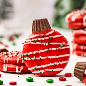 Red chocolate-covered Ritz cracker ornament cookie topped with a mini Reeseโs cup, drizzled with white chocolate and holiday sprinkles.