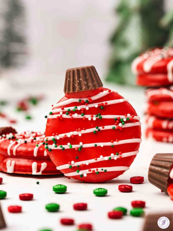 Red chocolate-covered Ritz cracker ornament cookie topped with a mini Reeseโs cup, drizzled with white chocolate and holiday sprinkles.
