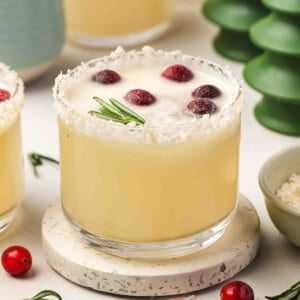 Glass of white Christmas punch with a coconut-rimmed edge, floating cranberries, and a fresh rosemary sprig.
