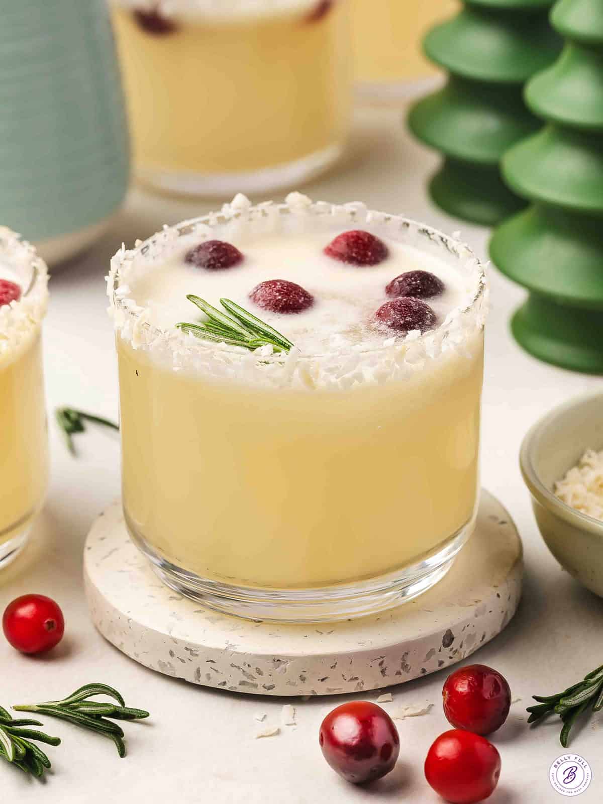 Glass of white Christmas punch with a coconut-rimmed edge, floating cranberries, and a fresh rosemary sprig.