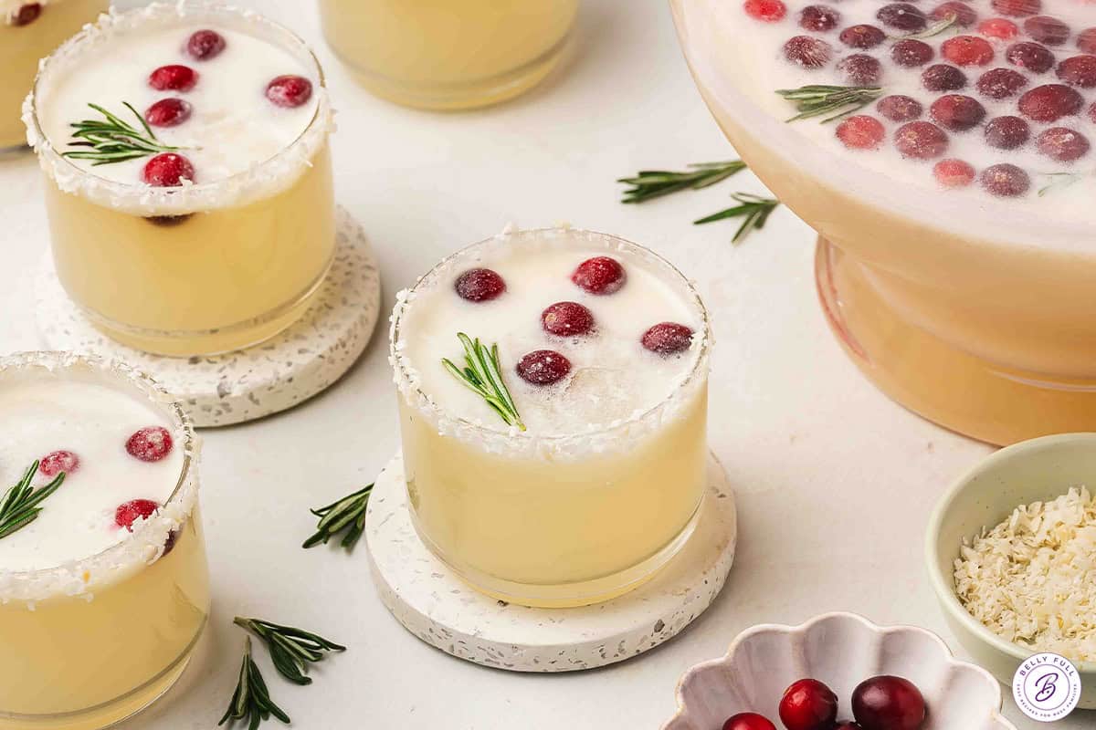 Glasses of white Christmas punch with coconut rims, cranberries, and rosemary arranged next to a full punch bowl.