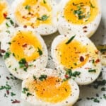 xHalved jammy eggs with soft golden yolks, seasoned with salt, pepper, and fresh herbs on a plate.