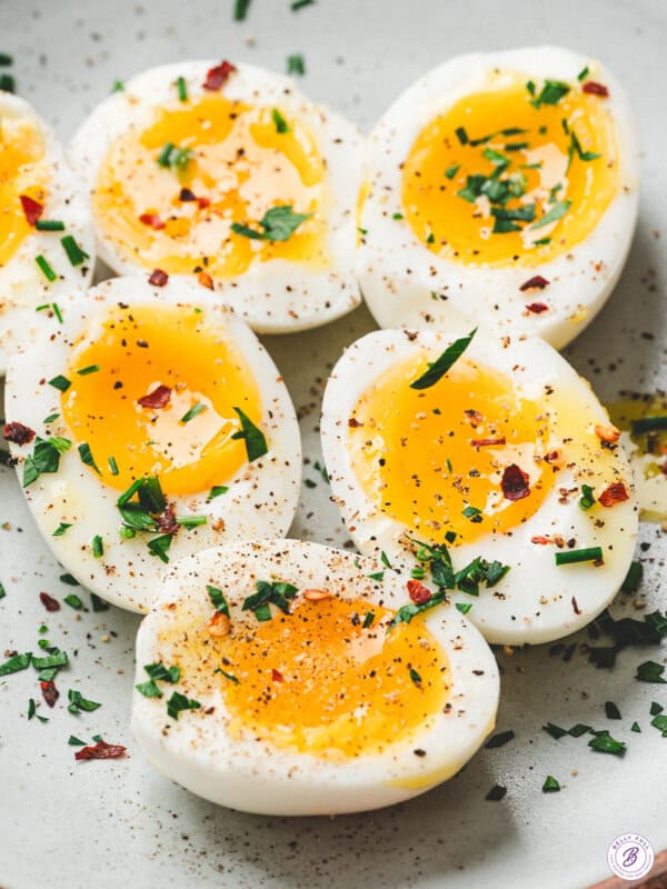 xHalved jammy eggs with soft golden yolks, seasoned with salt, pepper, and fresh herbs on a plate.