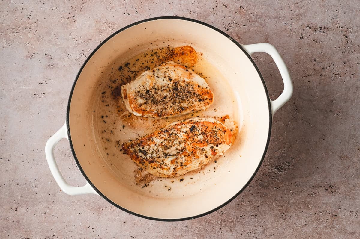 Seasoned chicken breasts browning in a Dutch oven with oil and herbs.