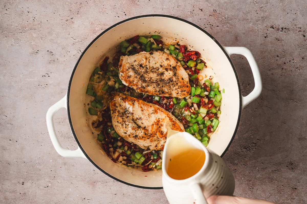 Pouring chicken broth over browned chicken, vegetables, and sun dried tomatoes in a Dutch oven.