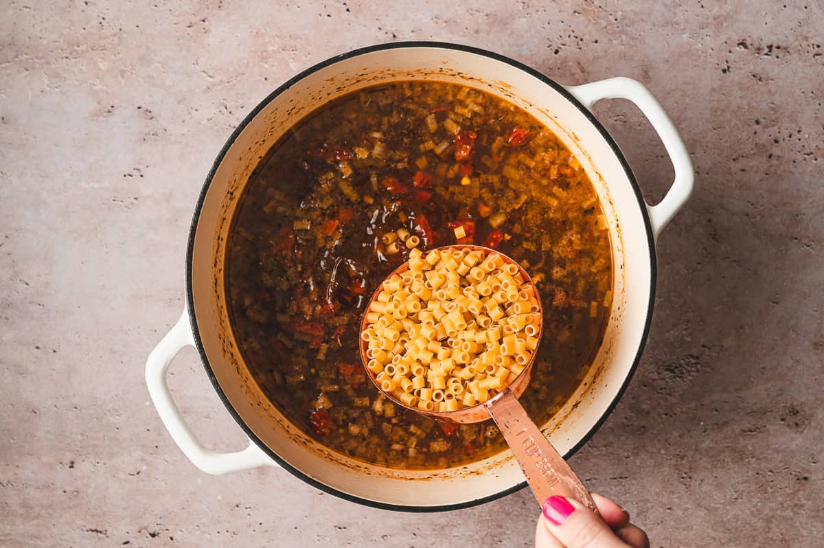 A measuring cup of dry ditalini pasta held over simmering soup broth in a pot.