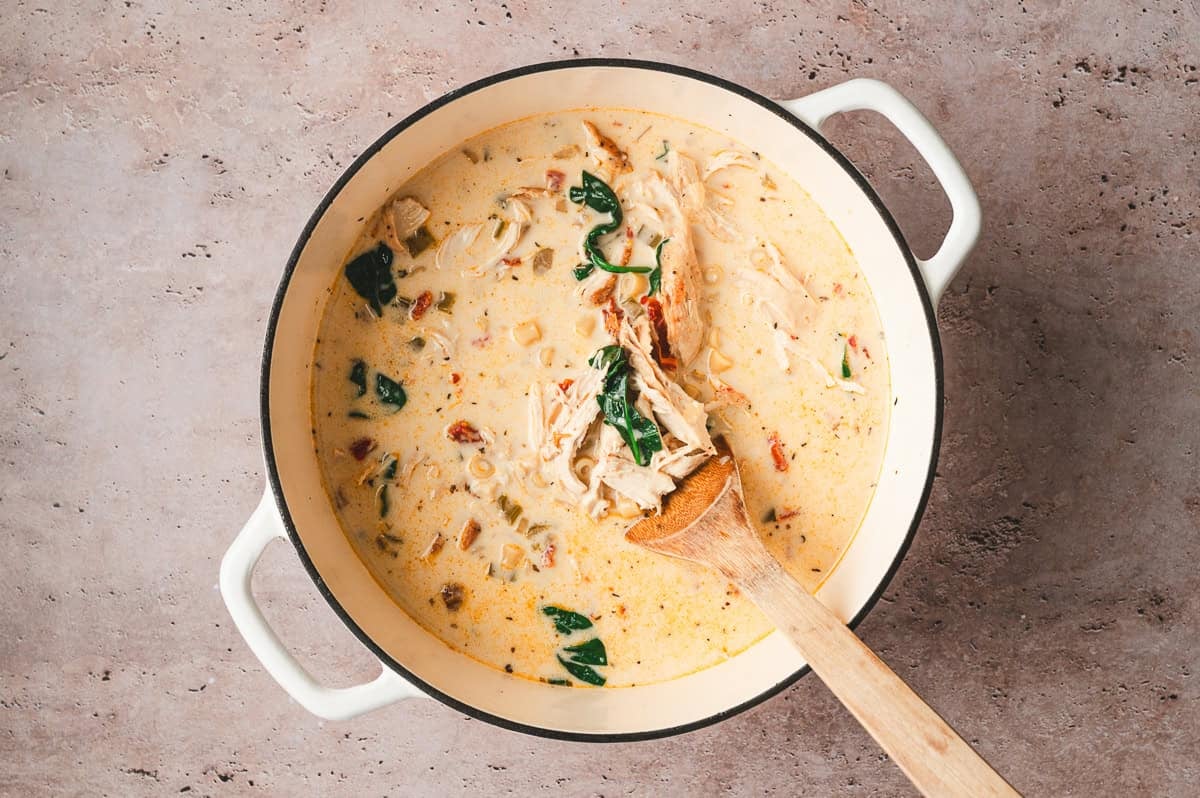 Creamy Marry Me Chicken Soup simmering in a Dutch oven with shredded chicken, spinach, sun dried tomatoes, celery, and pasta, stirred with a wooden spoon.