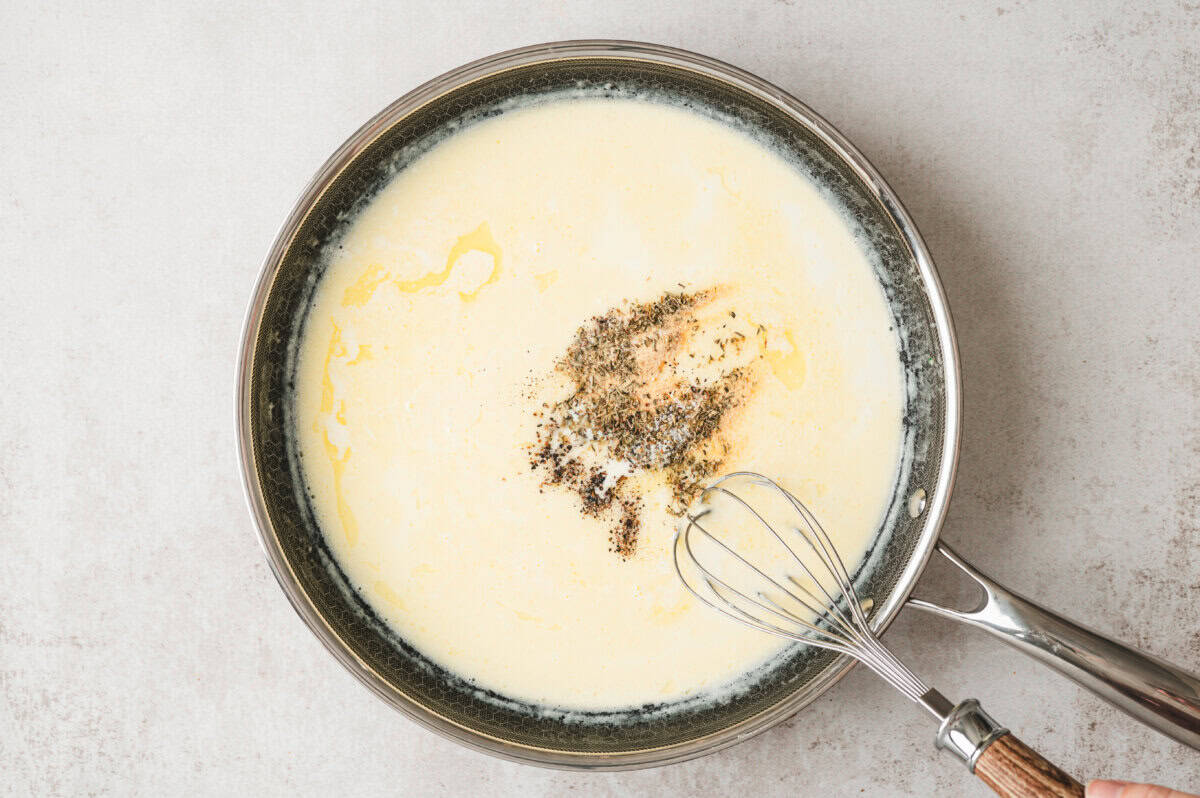Garlic, Italian seasoning, salt, and pepper being whisked into cream sauce in a skillet