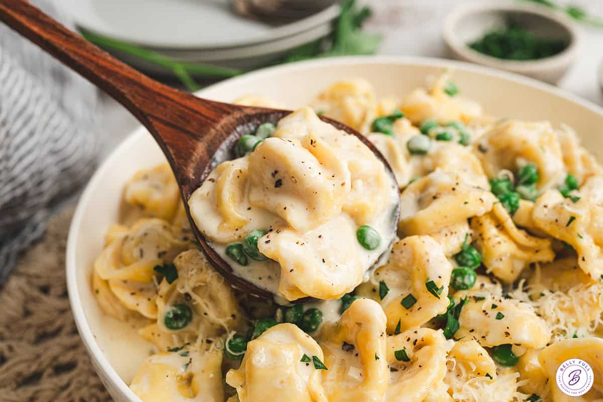 Spoon lifting cheese tortellini coated in creamy alfredo sauce with peas