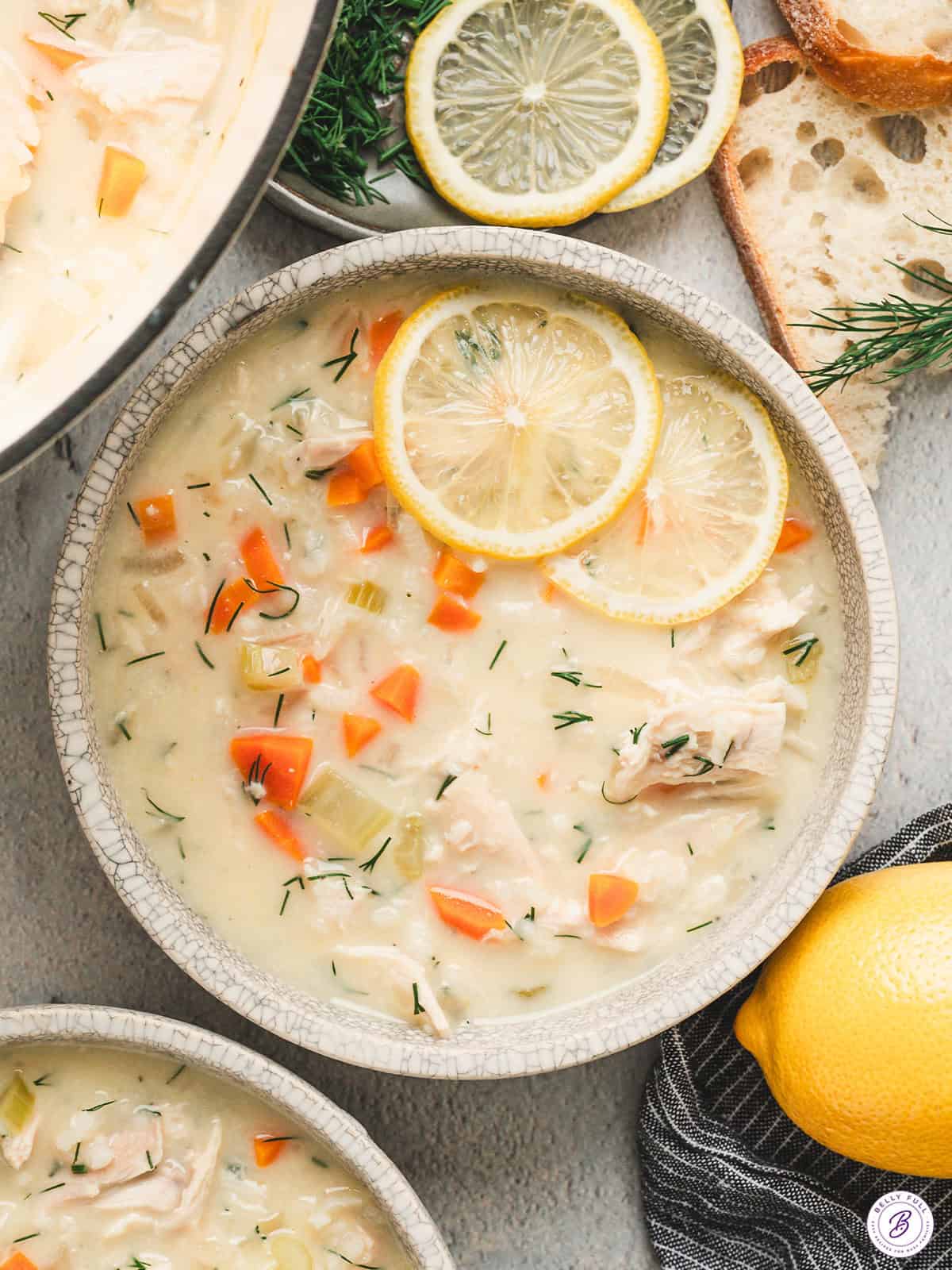 Creamy avgolemono soup with shredded chicken, rice, carrots, and dill, topped with lemon slices and served with crusty bread.