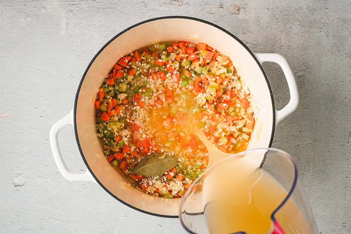 Chicken broth being poured into the pot with rice, vegetables, and a bay leaf for avgolemono soup.