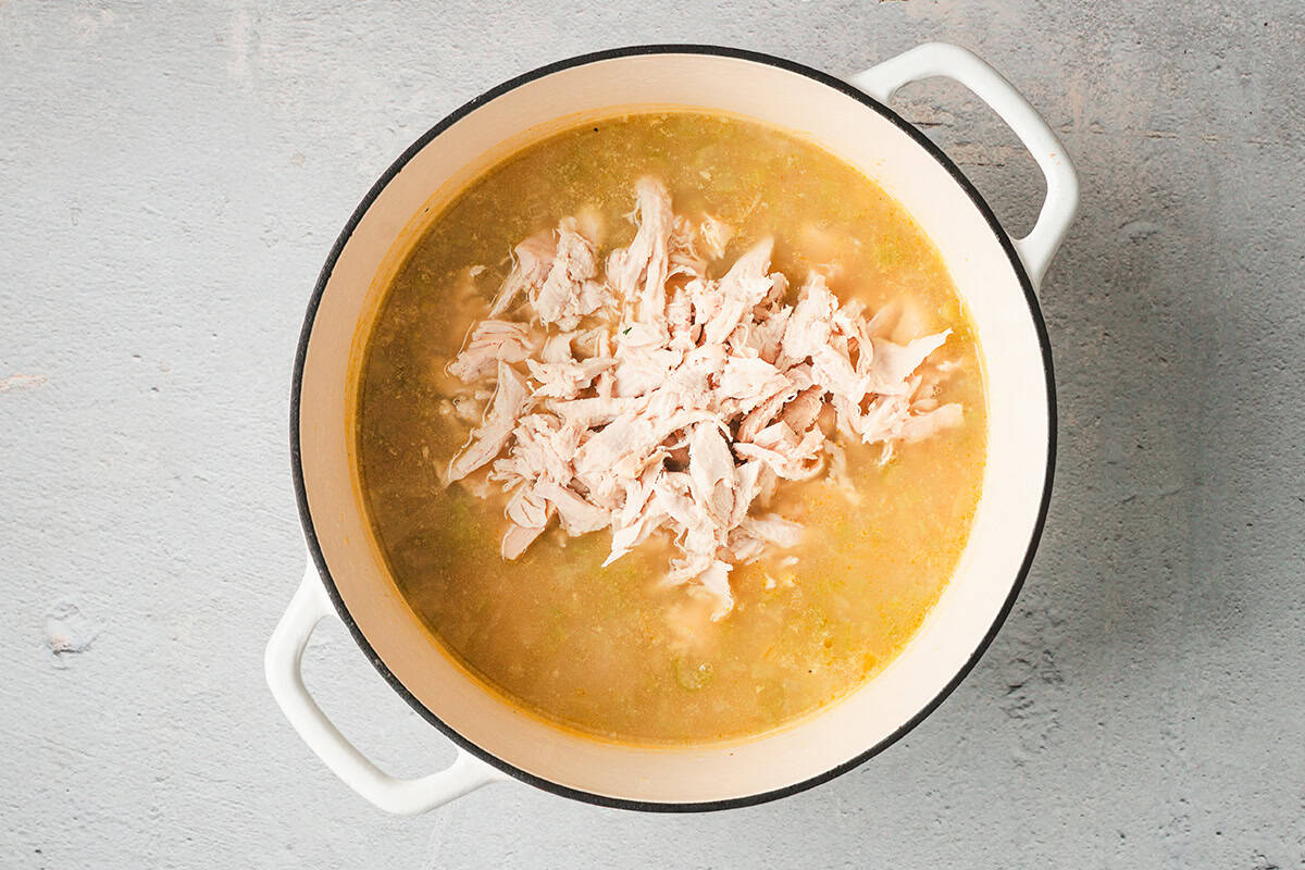 Shredded cooked chicken added to simmering chicken and rice soup in a large pot.