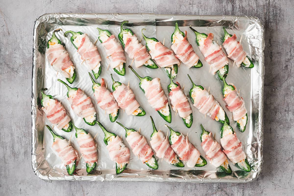 stuffed halved jalapeno peppers wrapped in raw bacon on baking sheet