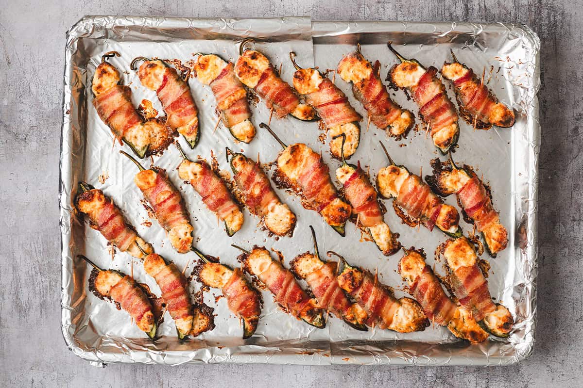 baked jalapeno peppers wrapped in bacon on baking sheet