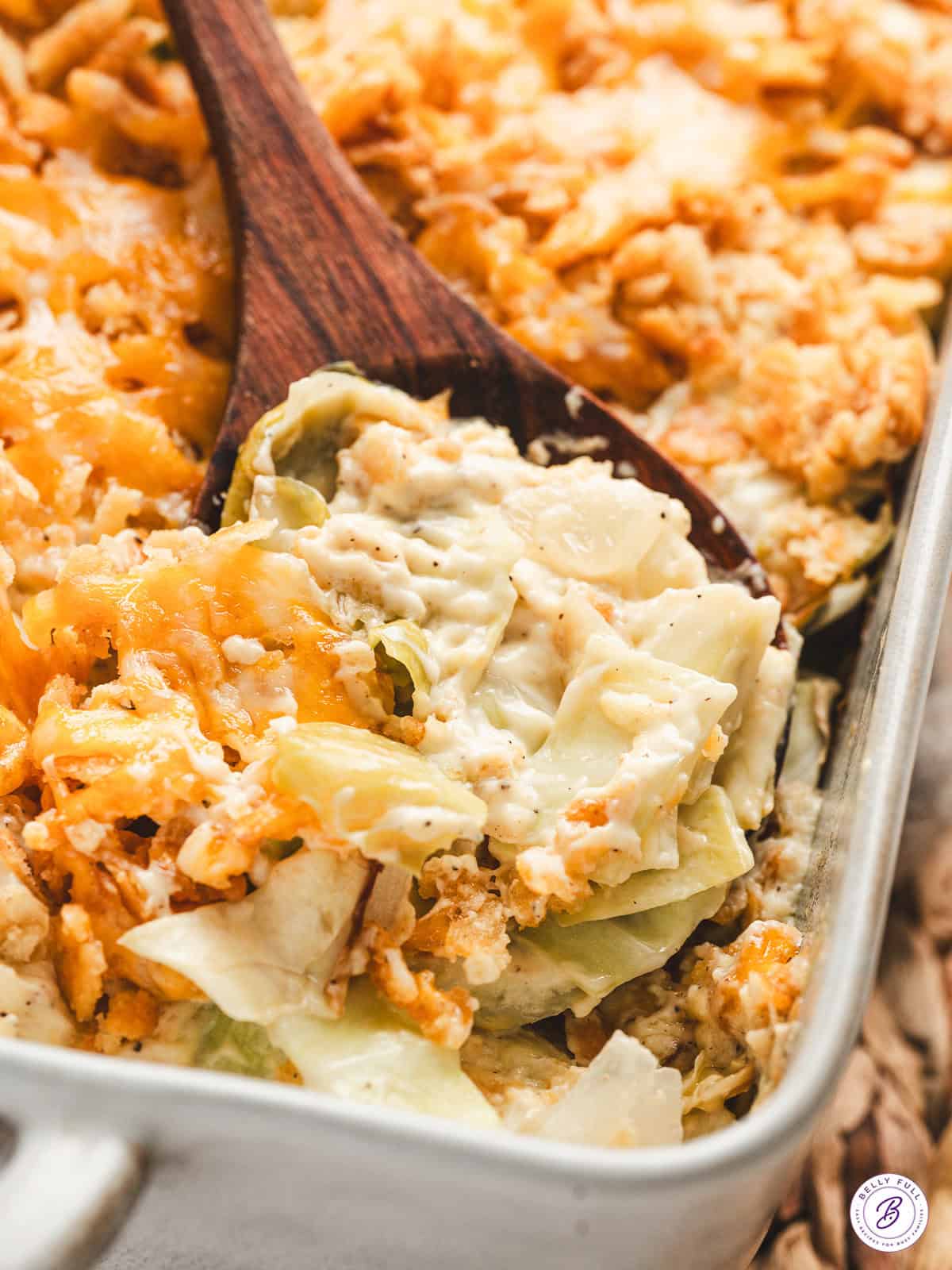 A wooden spoon scooping creamy cabbage casserole out of a baking dish.