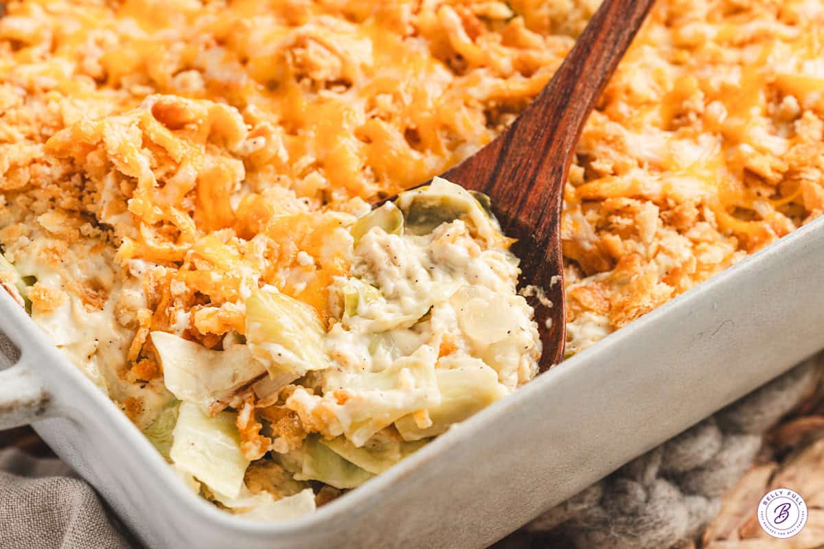 A wooden spoon scooping creamy cabbage casserole out of a baking dish.