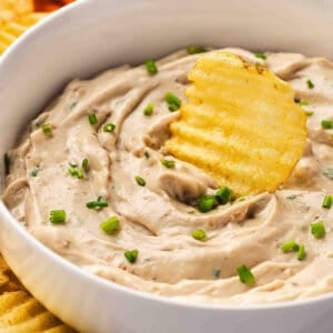French onion dip in a white bowl with chives, served with crinkle-cut potato chips and fresh vegetable sticks