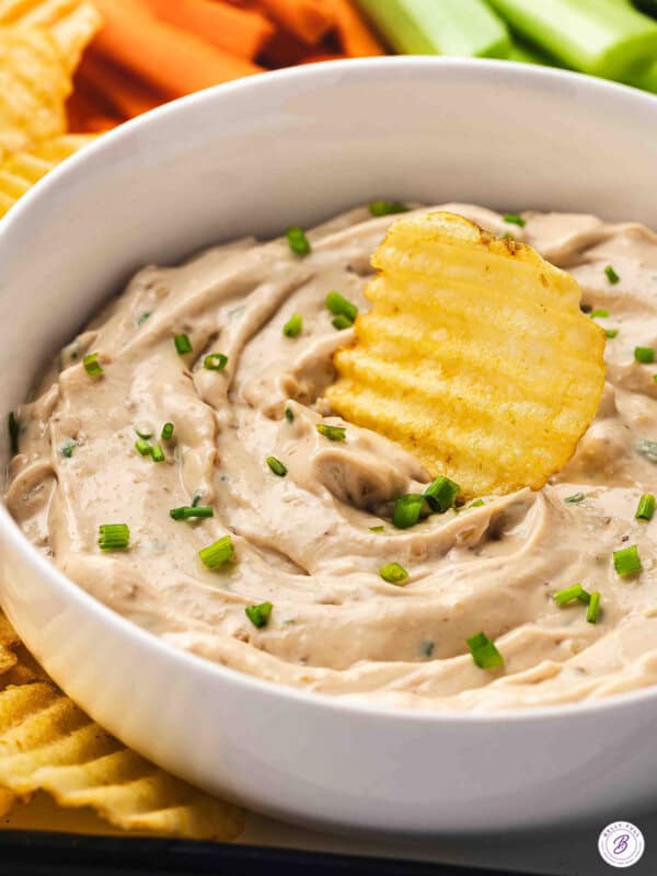 French onion dip in a white bowl with chives, served with crinkle-cut potato chips and fresh vegetable sticks