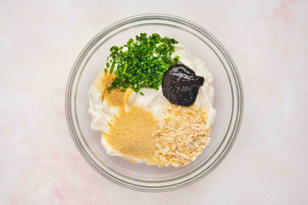 French onion dip ingredients in a glass bowl, including sour cream, dried onion flakes, chives, seasonings, and beef bouillon paste