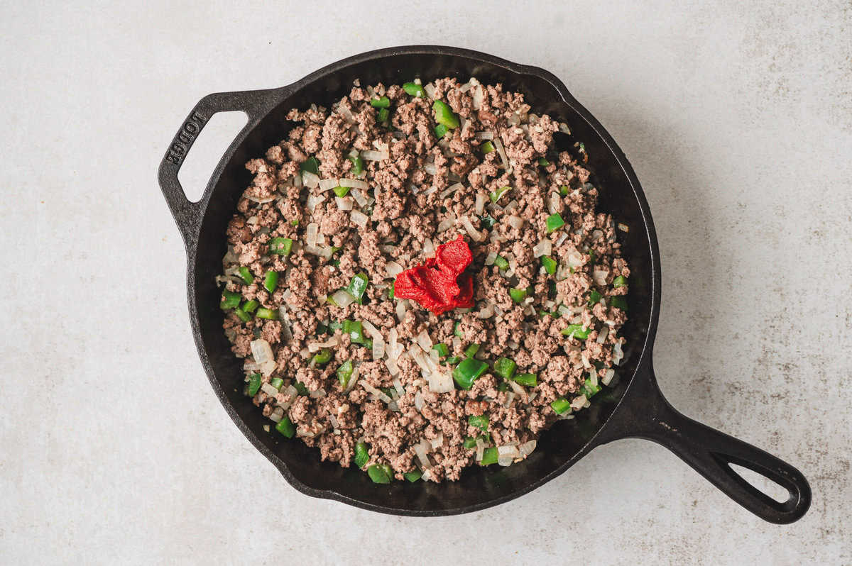 Tomato paste added to browned ground beef mixture in a cast-iron skillet, sitting on top before being stirred in.