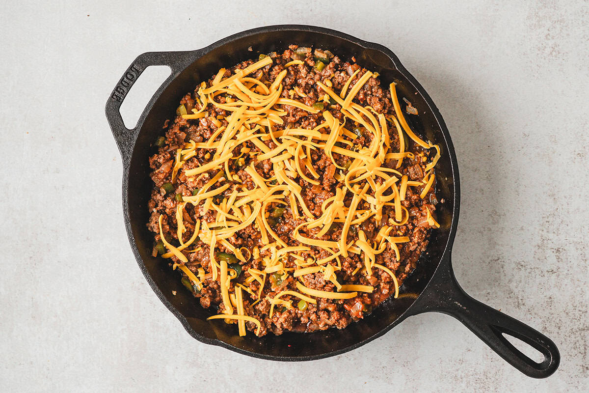 Shredded cheddar cheese sprinkled over the sloppy joe beef mixture in a cast-iron skillet before adding tater tots.