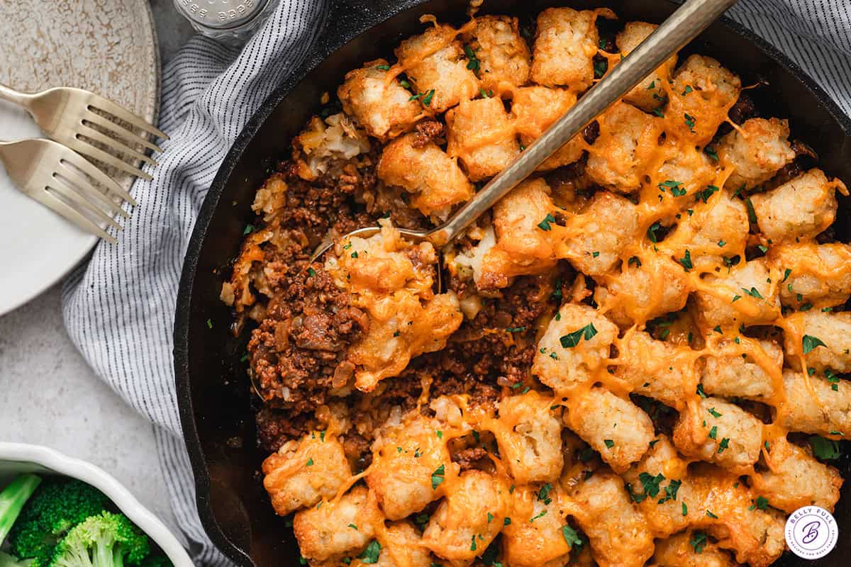 Spoon scooping sloppy joe tater tot skillet from a cast-iron pan, showing crispy tater tots, melted cheddar cheese, and the saucy ground beef filling underneath.