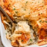 Close-up of au gratin potatoes being served from a white casserole dish with a golden, bubbly cheese topping and chopped parsley garnish.