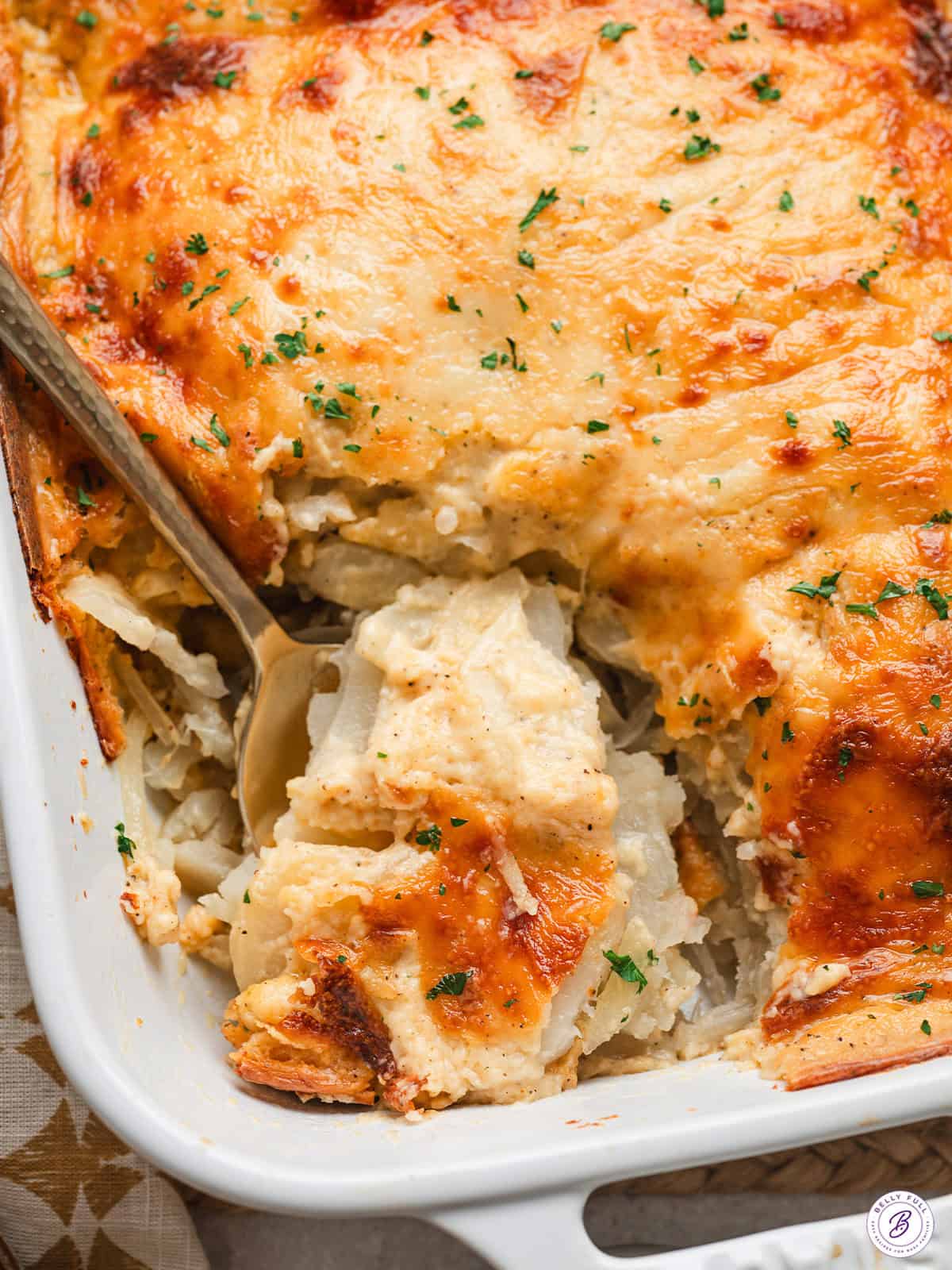 With thinly sliced potatoes, onions, and two types of cheese, these Au Gratin Potatoes are a must for every holiday. Close-up of au gratin potatoes being served from a white casserole dish with a golden, bubbly cheese topping and chopped parsley garnish.