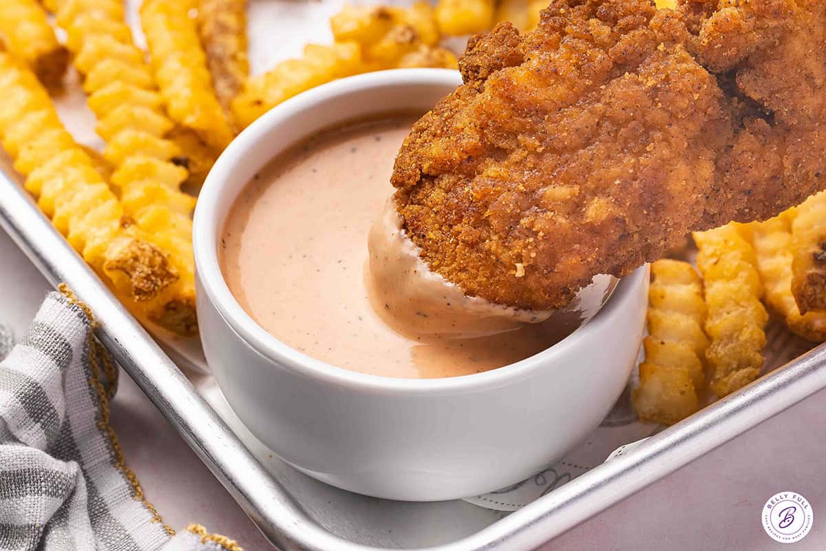 A chicken tender being dipped into a pot of homemade raising cane's sauce.