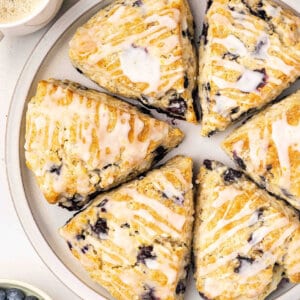 Lemon blueberry scones with lemon glaze arranged on a plate with fresh blueberries and coffee.