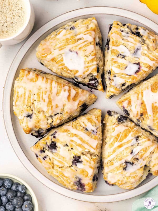 Lemon blueberry scones with lemon glaze arranged on a plate with fresh blueberries and coffee.