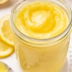 Jar of homemade lemon curd with a glossy swirl on top, surrounded by fresh lemon slices.