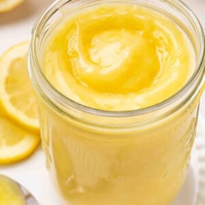 Jar of homemade lemon curd with a glossy swirl on top, surrounded by fresh lemon slices.