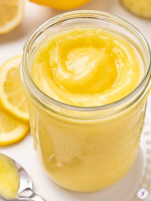 Jar of homemade lemon curd with a glossy swirl on top, surrounded by fresh lemon slices.