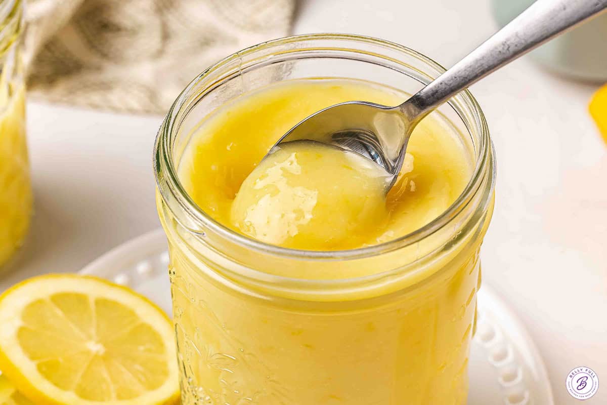 Spoon scooping creamy lemon curd from a mason jar with lemon slices in the background.
