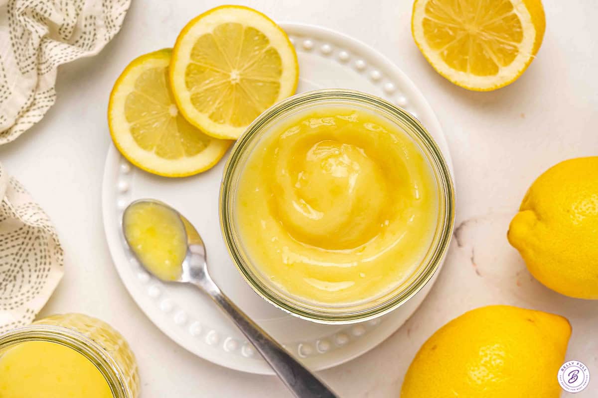Overhead view of homemade lemon curd in a jar on a plate with fresh lemons and a spoon.