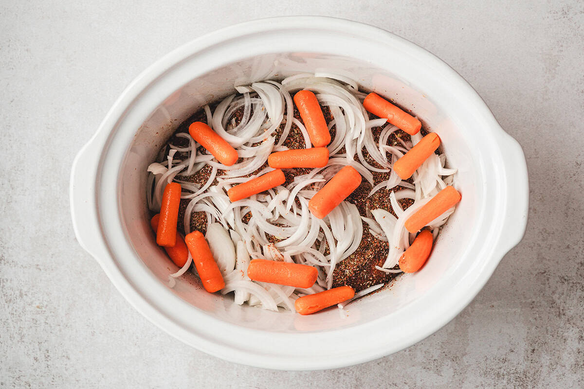 Sliced onions and baby carrots layered over seasoned brisket in a slow cooker before cooking