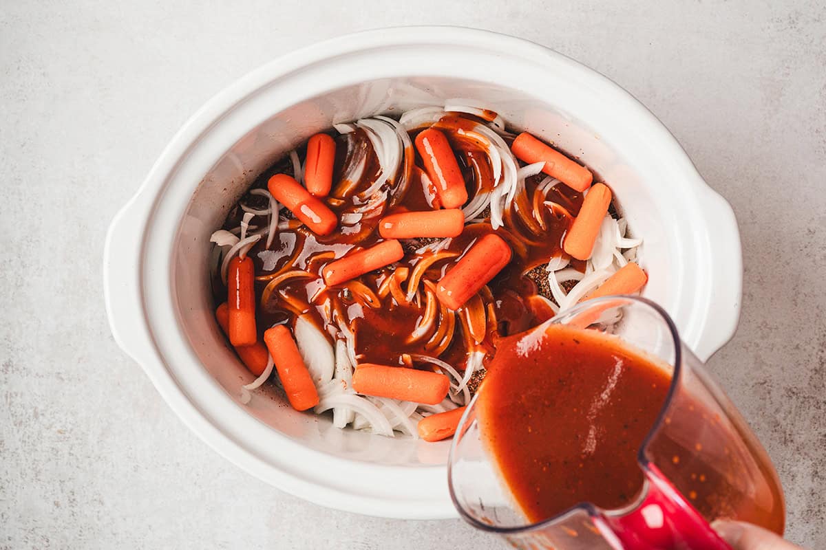 Barbecue sauce mixture being poured over brisket, onions, and carrots in a slow cooker