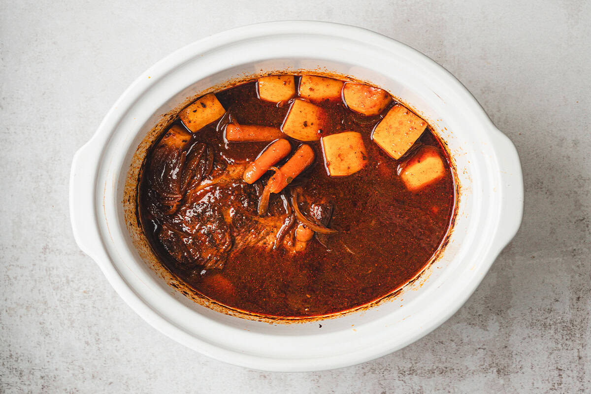 Cooked brisket in slow cooker with carrots and potatoes fully submerged in rich barbecue sauce