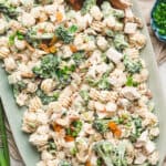 Chicken broccoli pasta salad served on a large platter with creamy dressing, broccoli florets, diced chicken, raisins, almonds, and scallions topped with wooden serving spoons