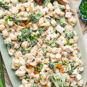 Chicken broccoli pasta salad served on a large platter with creamy dressing, broccoli florets, diced chicken, raisins, almonds, and scallions topped with wooden serving spoons