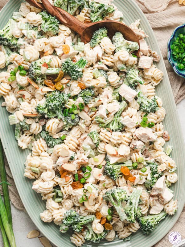Chicken broccoli pasta salad served on a large platter with creamy dressing, broccoli florets, diced chicken, raisins, almonds, and scallions topped with wooden serving spoons