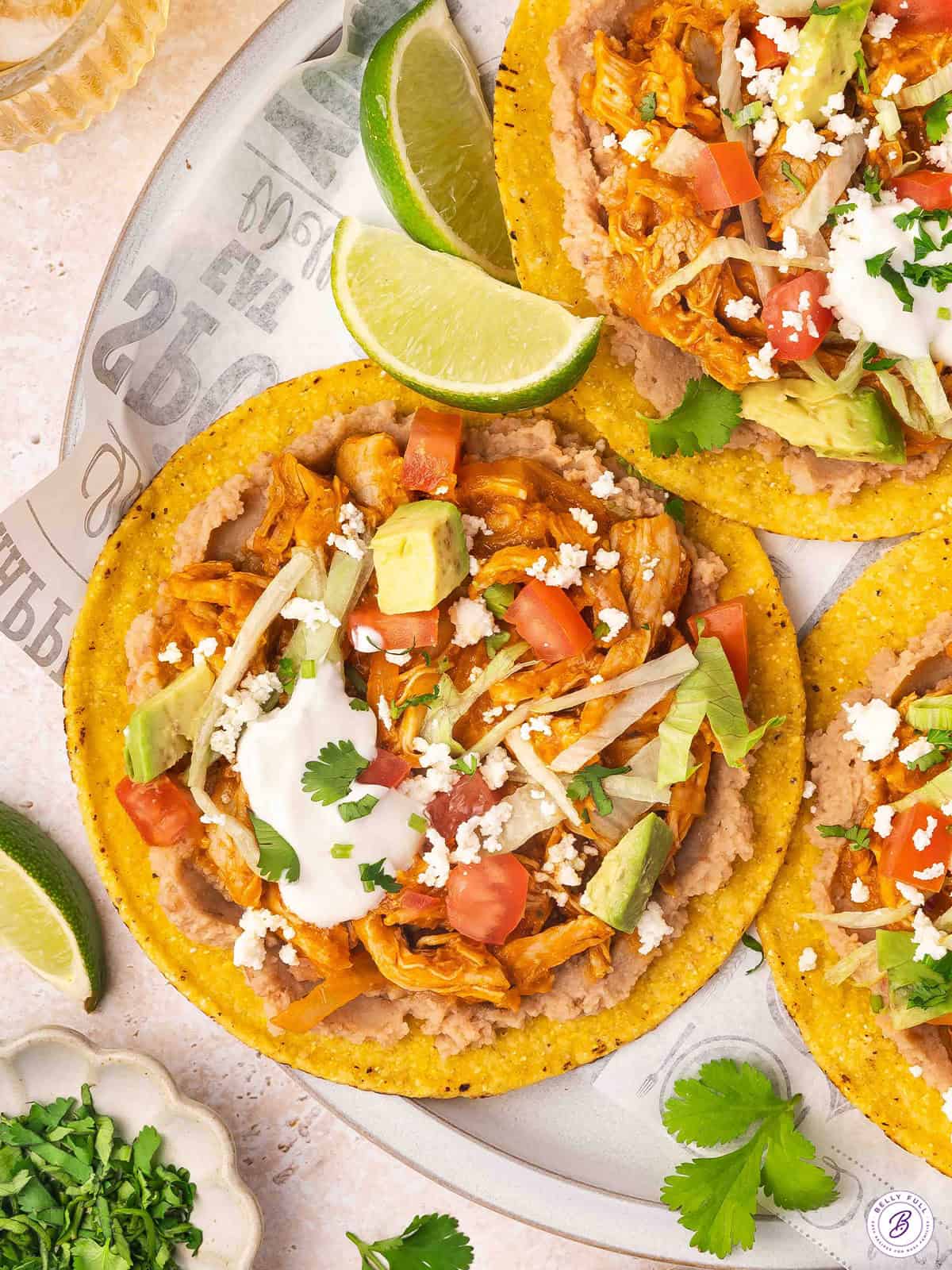 Overhead view of chicken tinga tostadas on a plate with cheese, sour cream and lime wedge toppings.