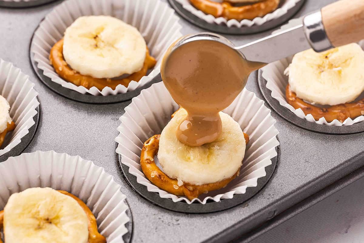 A spoon drizzling peanut butter on a slice of banana butter and pretzel in a muffin case.