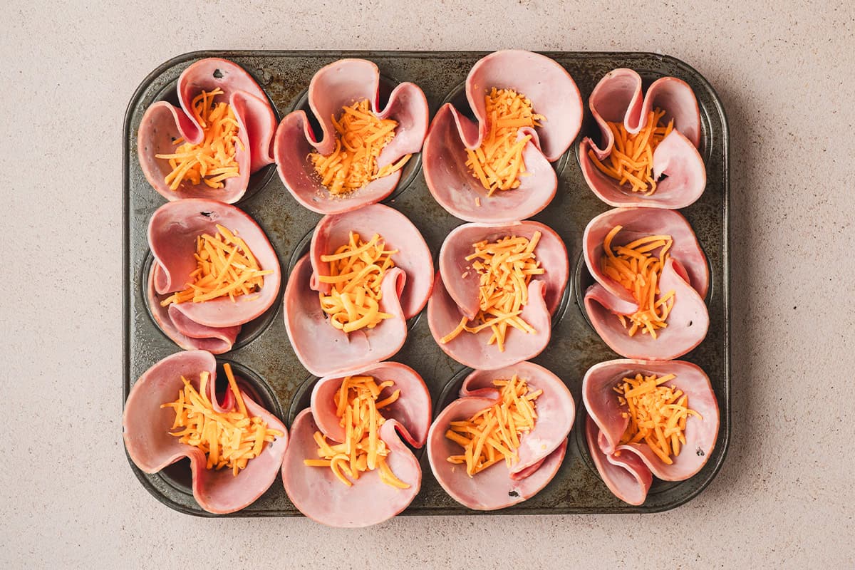 Ham placed in a muffin pan, with shredded cheese placed on top.