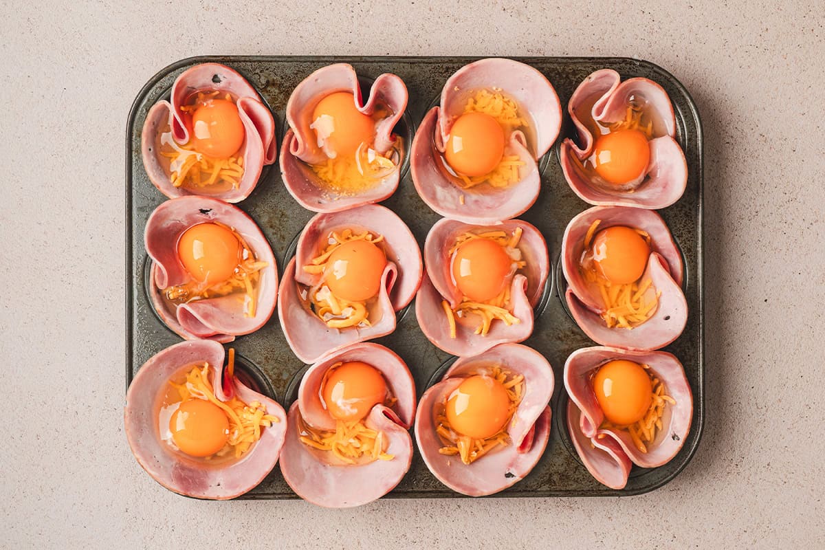 Ham placed in a muffin pan, with shredded cheese and eggs cracked on top.