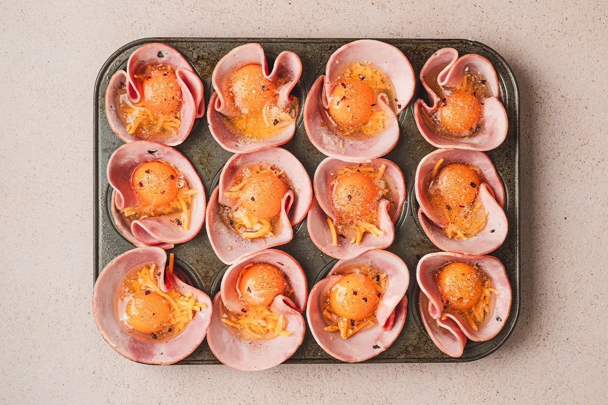 Ham placed in a muffin pan, with shredded cheese and eggs cracked on top with seasonings sprinkled over them.